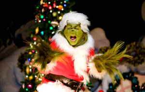 A Man Dressed As The Grinch Is Performing In Front Of A Christmas Tree Wallpaper