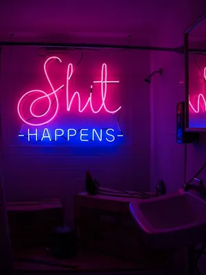 A Neon Sign That Says Shit Happens In A Bathroom Wallpaper