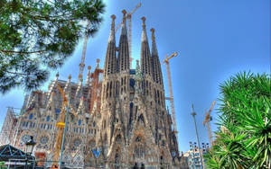 Majestic Sagrada Familia Surrounded By Lush Trees And Industrial Cranes Wallpaper