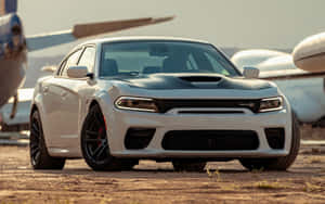 The White Dodge Charger Srt Parked Next To An Airplane Wallpaper